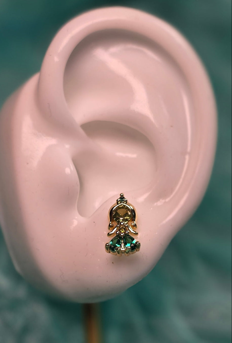 Gold earring with a green gemstone on a white mannequin ear against a teal background